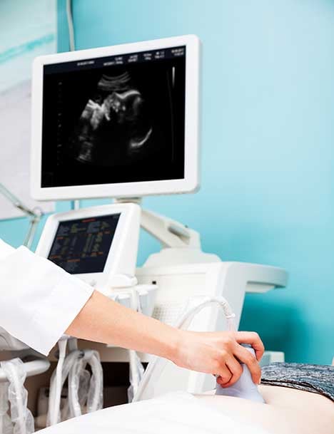 Healthcare professional performing an ultrasound exam on a patient while a fetal ultrasound image is displayed on the monitor in a clinical setting.