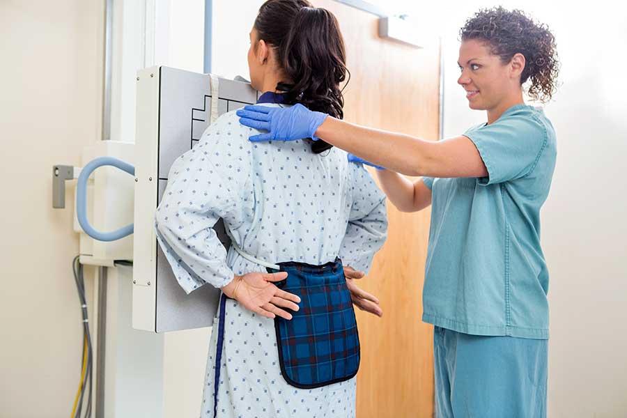 Radiologic technologist positioning a patient for a chest X-ray exam. The patient stands against the imaging plate wearing a hospital gown and protective lead apron while the technologist adjusts their posture.